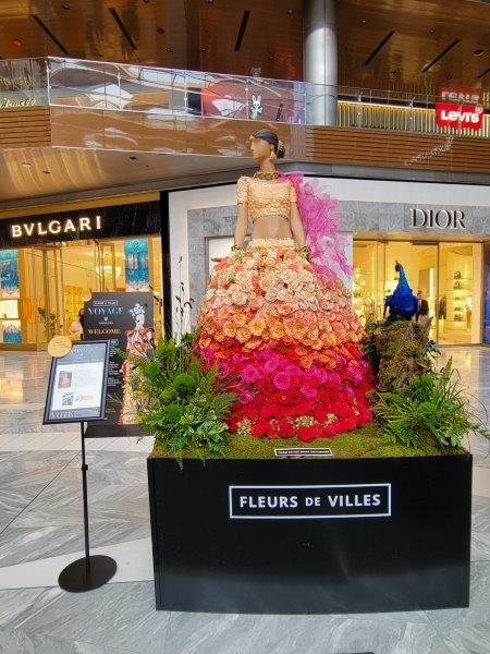 India floral mannequin in a shaded saree with flowers running from peach to pink to red, and positioned next to a floral blue peacock