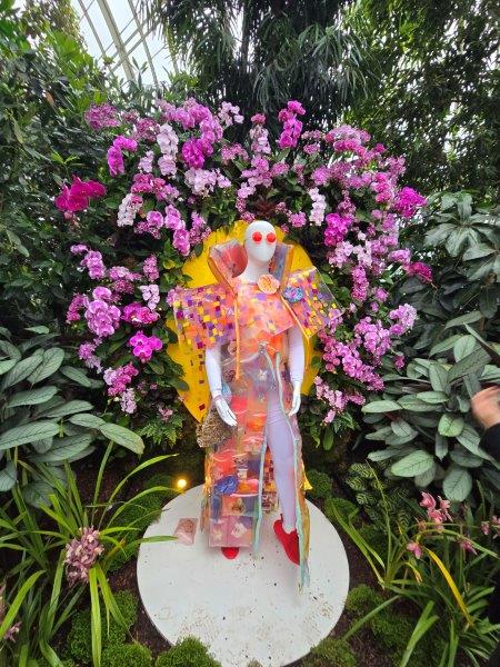 Mannequin with an orange dress with sewn in orchids framed by pink orchides