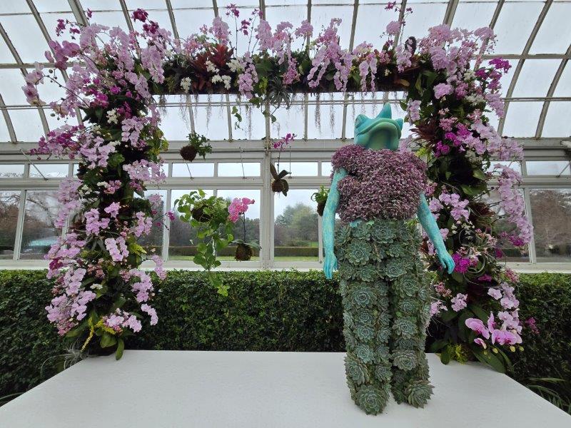 Frog mannequin on a platform wearing a green orchid pants with a light pink orchid frame as a backdrop 